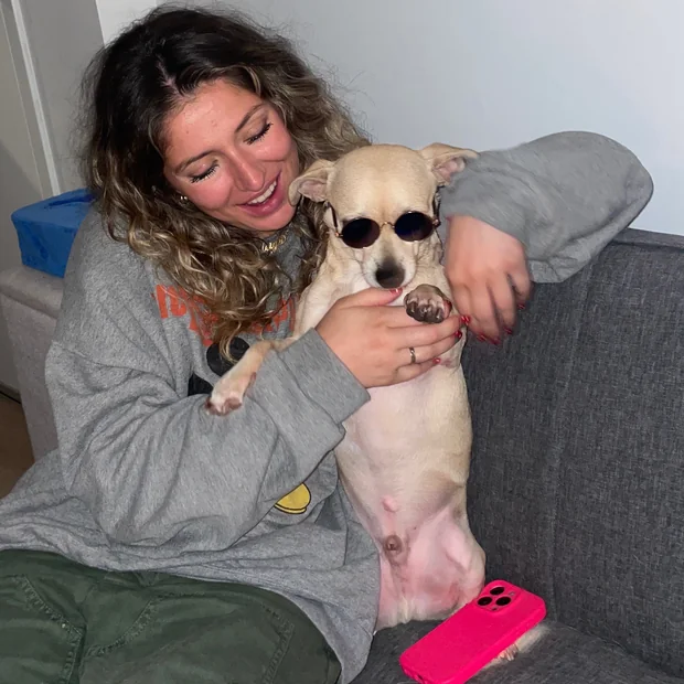 Karleigh on a sofa, laughing, with Noodle — a small tan chihuahua in round dark sunglasses — propped up beside her.